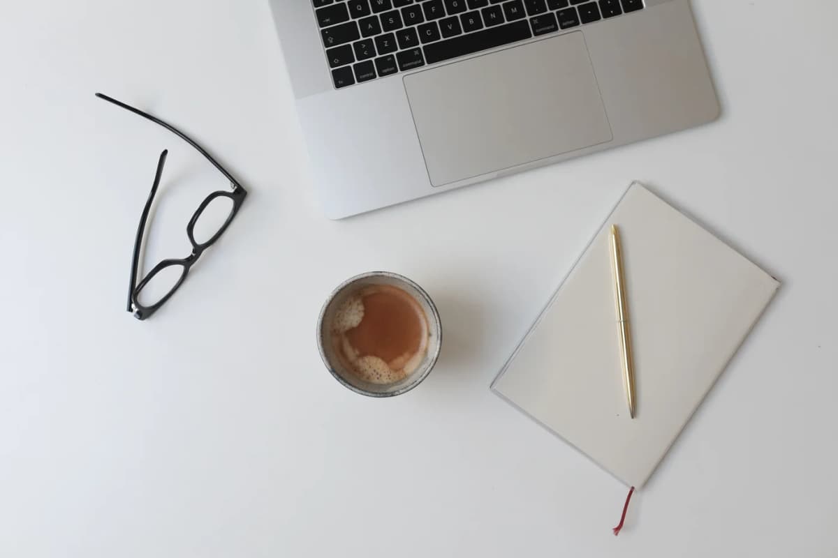 Laptop keyboard, glasses, coffee and a notebook on a white desk