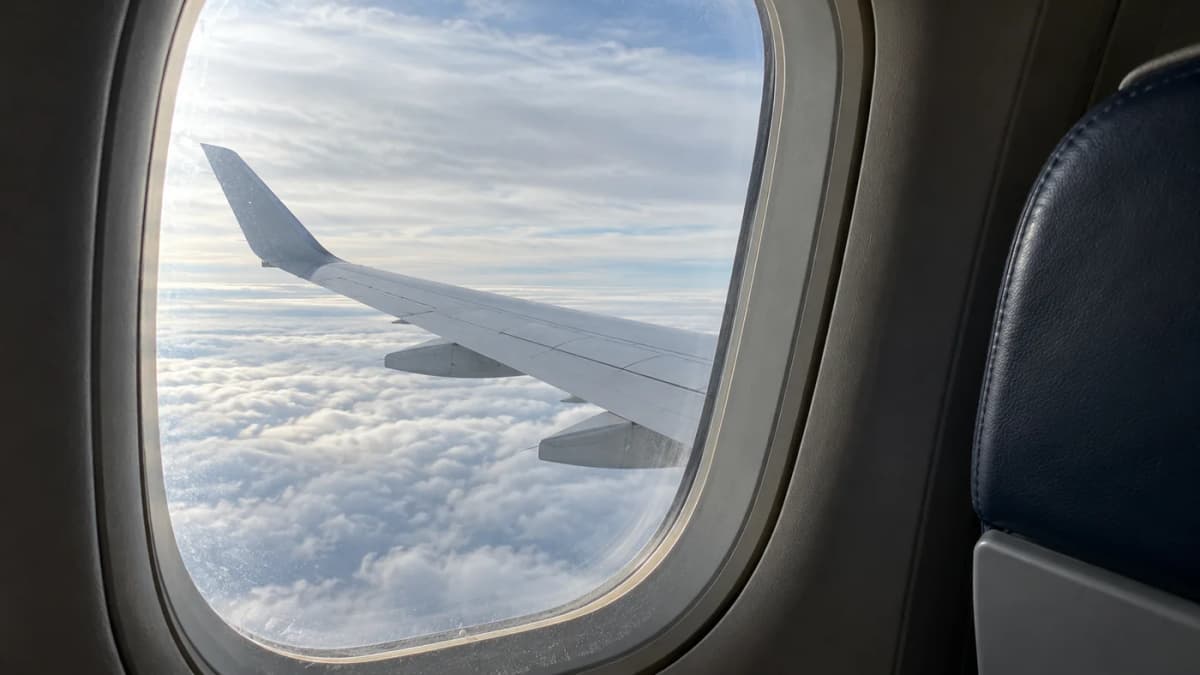 An unbranded aircraft wing seen through an economy-cabin window above clouds.